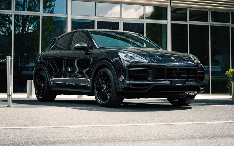 Porsche Cayenne Turbo GT noir vu de trois quarts avant, à l'arrêt devant une façade vitrée, en plein soleil.
